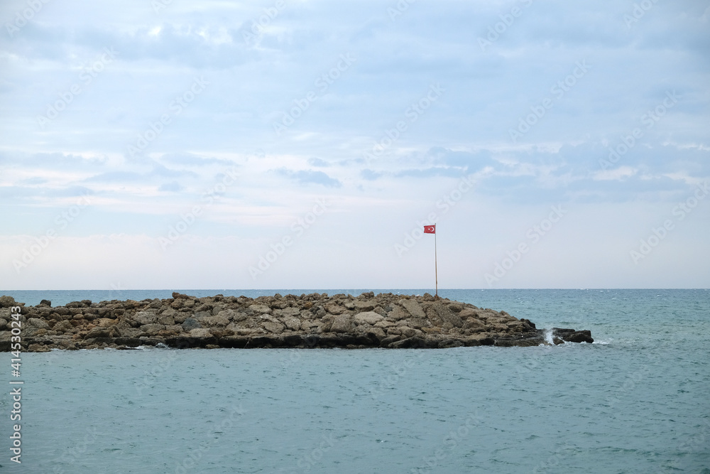 Landscape with open Mediterranean sea and the entrance to the marina with Turkish flag on flagpole on the edge of the coast in the overcast day