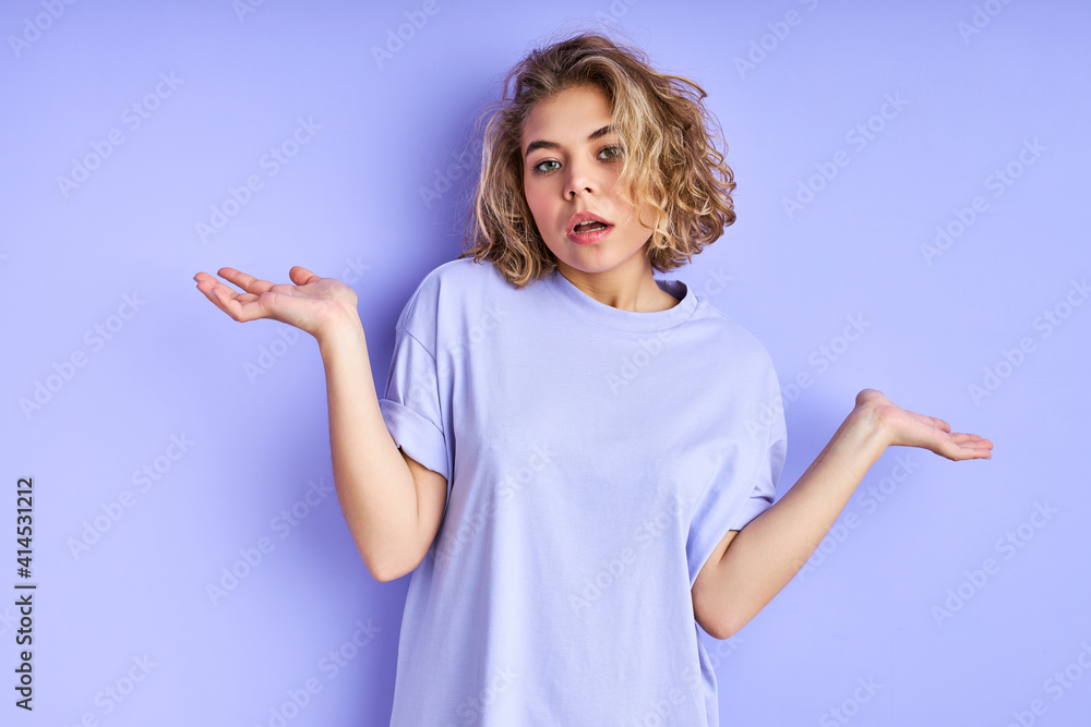 Fototapeta premium unsure female model shrugs shoulders with uncertainty isolated in studio, makes questioning gesture, has clueless and confused look, poses alone against purple wall. Who cares