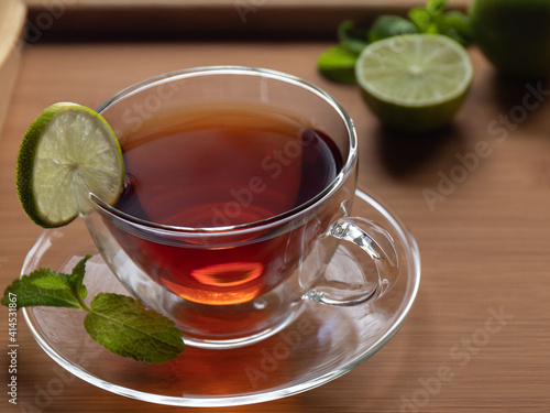 Black tea with a sprig of mint and a slice of lime in a transparent cup with a saucer in focus on a wooden tray. Lime and mint in the background in a blur. The concept of the menu for restaurants