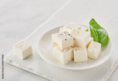 Bowl of diced soft cheese feta with basil leaf on white plate.