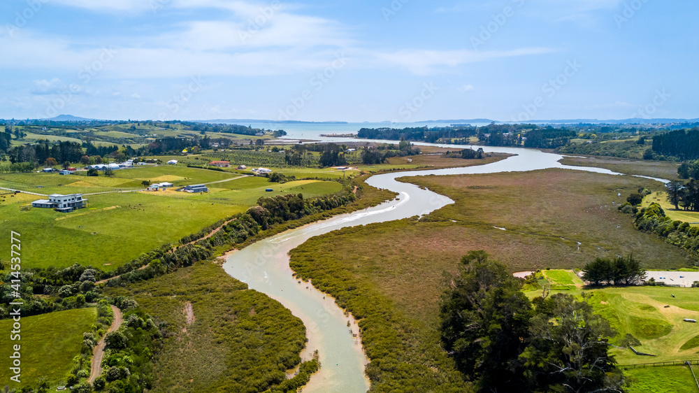 Aerial view of a small river running to the sea through green countryside spotted with little farms. Auckland, New Zealand.