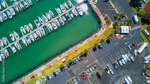 Aerial view on a marina with boat and yachts resting on calm water and cars parked along the shore on a busy plaza. Auckland, New Zealand.