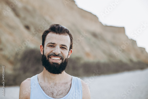 portrait of a stylish young man with a beard and a face on which the sea sand on the background of the sea shore