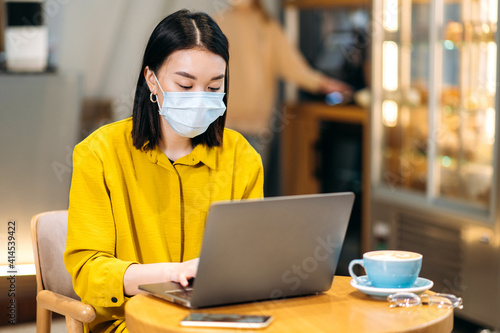 Female successful freelancer or student using a laptop for remote work or distance learning sits at the cafeteria with a cup of coffee. Asian girl wearing medical mask answers emails,browsing internet