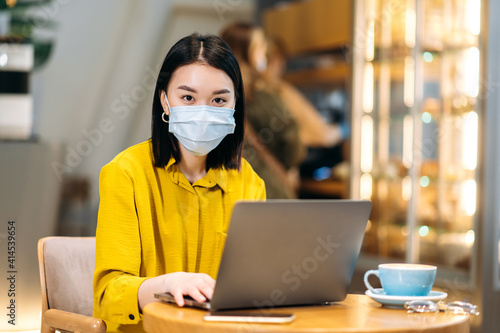 Asian female freelancer or student using a laptop for remote work or distance learning sits at the cafeteria with a cup of coffee, wearing medical mask answers emails,browsing internet