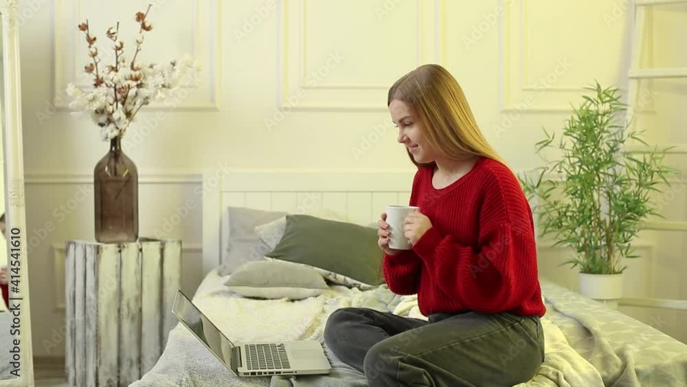Beautiful young woman working on a laptop sitting on the bed at home, she is drinking coffee and smiling. Work from home during quarantine. Girl in a sweater and jeans at home on the bed Beautiful young woman working on a laptop sitting on the bed at home, she is drinking coffee and smiling. Work from home during quarantine. Girl in a sweater and jeans at home on the bed