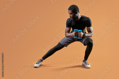 Fototapeta Naklejka Na Ścianę i Meble -  athletic man lunges with dumbbells in hands, do sportive exercises, lead healthy lifestyle isolated on brown background