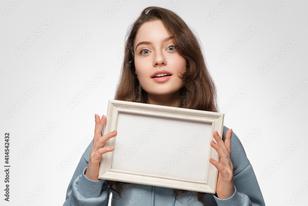 Young woman with an empty picture frame horizontally in her hand on a light blue background. The frame has a space for your text or photo. Light blue hoodie, blue eyes long hair. Young woman with an empty picture frame horizontally in her hand on a light blue background. The frame has a space for your text or photo. Light blue hoodie, blue eyes long hair.