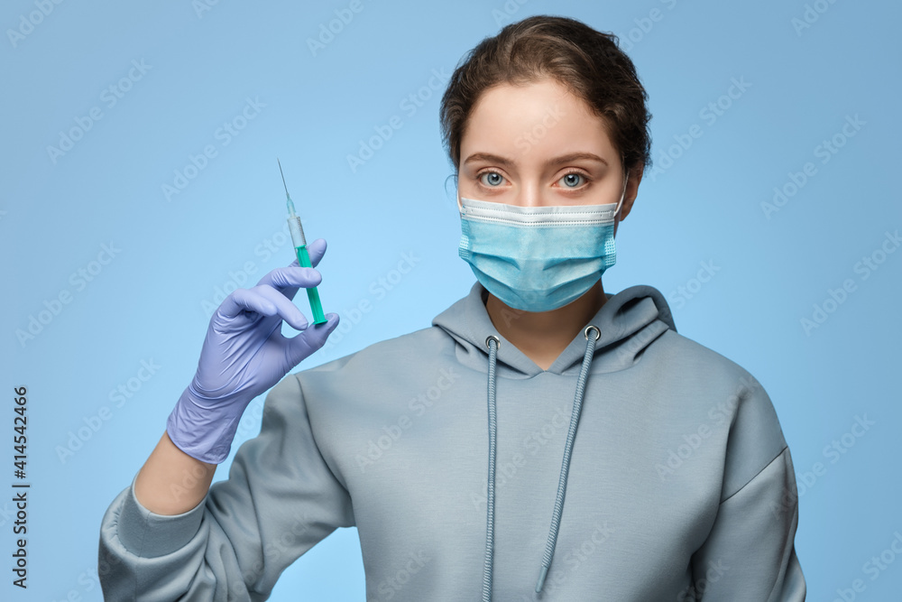 A young woman in a medical mask holds a syringe in her hand, blue latex gloves on her hand. The nurse prepares for the injection. A young woman in a medical mask holds a syringe in her hand, blue latex gloves on her hand. The nurse prepares for the injection.