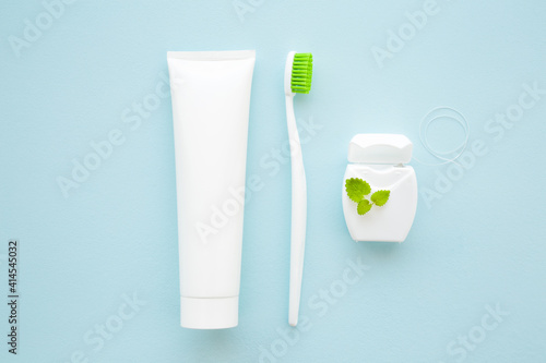 Toothbrush with green bristles, white tube of toothpaste, container of dental floss and peppermint leaf on light blue table background. Pastel color. Closeup. Top down view. People teeth hygiene.