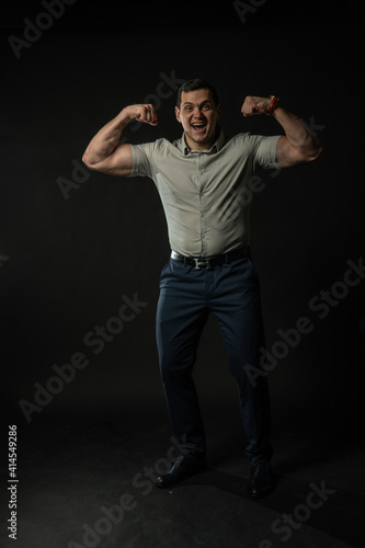 Shows off muscle and smiling bodybuilder in grey shirt on black background confident elegance model, portrait modern suit, studio beautiful. Content boy, shirt cool black © Ilya