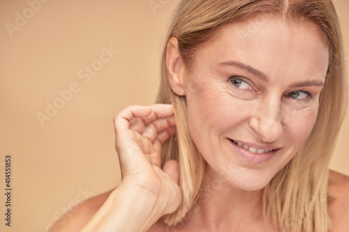 Feeling attractive. Portrait of a beautiful middle aged woman touching her hair, looking aside and smiling while posing over beige background