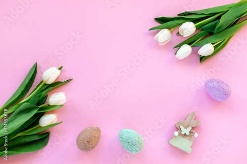 Easter festive background with painted eggs, wooden toy rabbit and white tulips on pink. Top view, flat lay, copy space