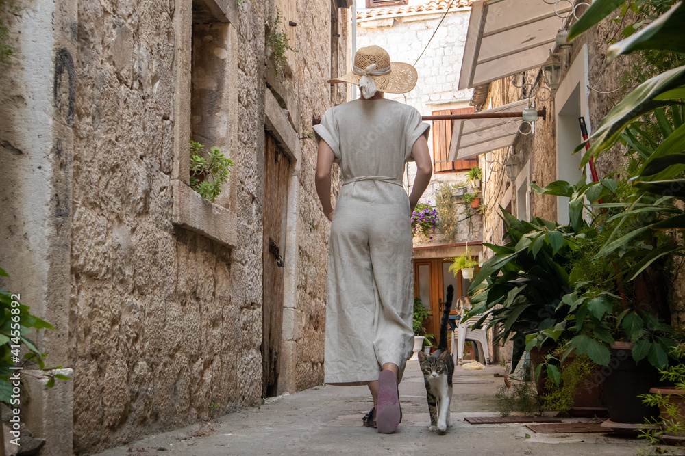 Rear view of beautiful blonde young female traveler wearing straw sun hat sightseeing and enjoying summer vacation in an old traditional costal town at Adriatic cost, Croatia.