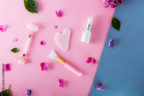 Top view composition with pink jade roller massager, gouache scraper, applying brush and skin serum on pink and blue background with hydrangea flower petals. Anti-age, lifting, toning skin treatment