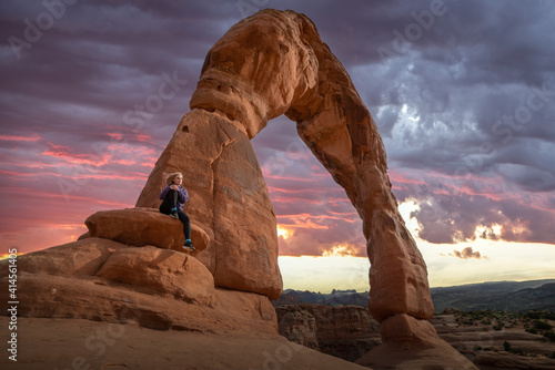 A alone, isolated woman tourist sitting beside Delicate Arch in National Park, Utah during summer time with pink, purple sunset in background and wind blowing in her face.  photo