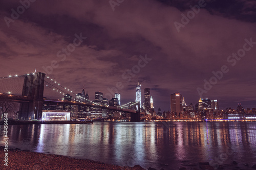 Illuminated Brooklyn Bridge And Buildings By River Against Sky At Night in New York City