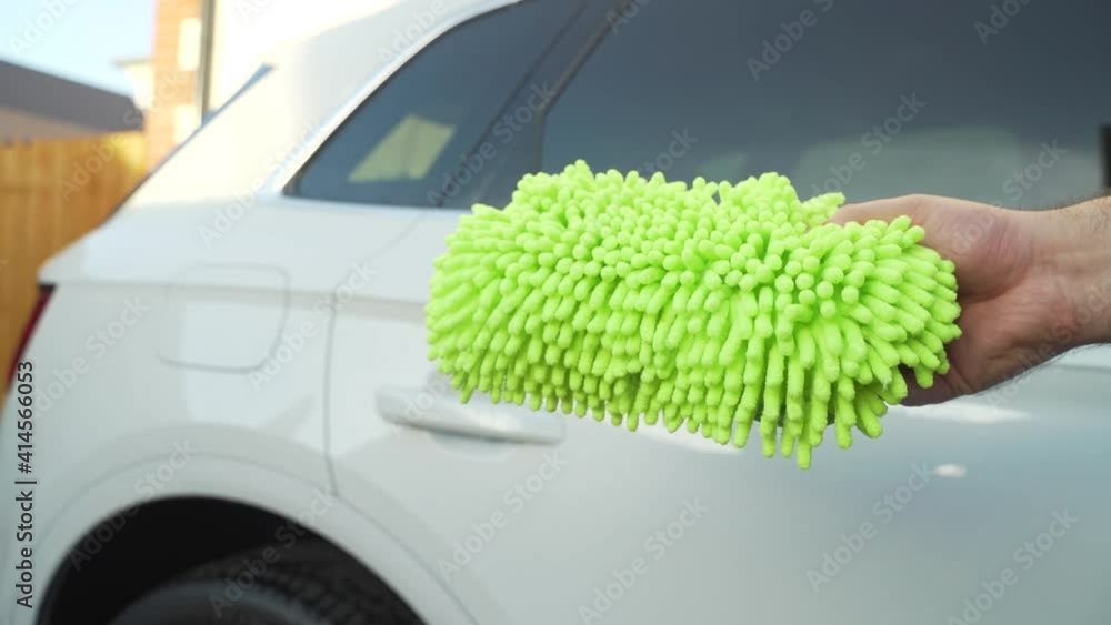 Product Demonstration. Man shows a bright green-yellow microfiber noodle sponge to the camera which will be used to wash the white premium SUV in the background. Product Demonstration. Man shows a bright green-yellow microfiber noodle sponge to the camera which will be used to wash the white premium SUV in the background.