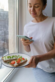 Woman takes a photo on smart phone, holding plate with rye crisp bread with creamy vegetarian cheese tofu, cherry tomato and rucola micro greens. Healthy food , tasty breakfast, gluten free, diet. 