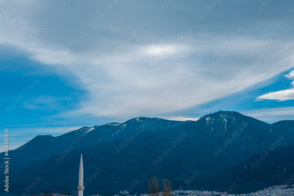 Magnificent ulu mountain (uludag) view from the city center together with minaret of a mosque and dried trees. Huge clouds and snow remains on top of the mountain.