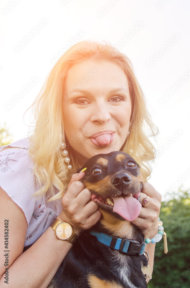 A beautiful blonde girl in a light dress and a cute dog have fun together and show their tongues.  Vertical view