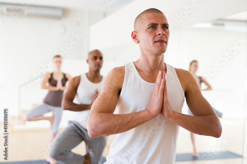 Concentrated man standing on one leg in Eka Pada Pranamasana pose during group yoga training in gym