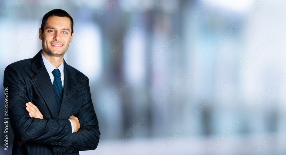 Portrait of happy confident businessman in black suit and blue tie, with crossed arms pose, copy space for some text, advertising or slogan, standing against blured office background Portrait of happy confident businessman in black suit and blue tie, with crossed arms pose, copy space for some text, advertising or slogan, standing against blured office background