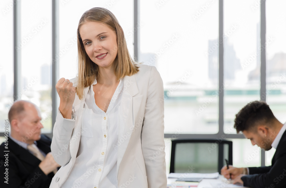 Adults caucasian woman in white shirt and blue jean standing smile fist hand and look at a camera feel confident which man staff and boss in the blur background do working hard Adults caucasian woman in white shirt and blue jean standing smile fist hand and look at a camera feel confident which man staff and boss in the blur background do working hard