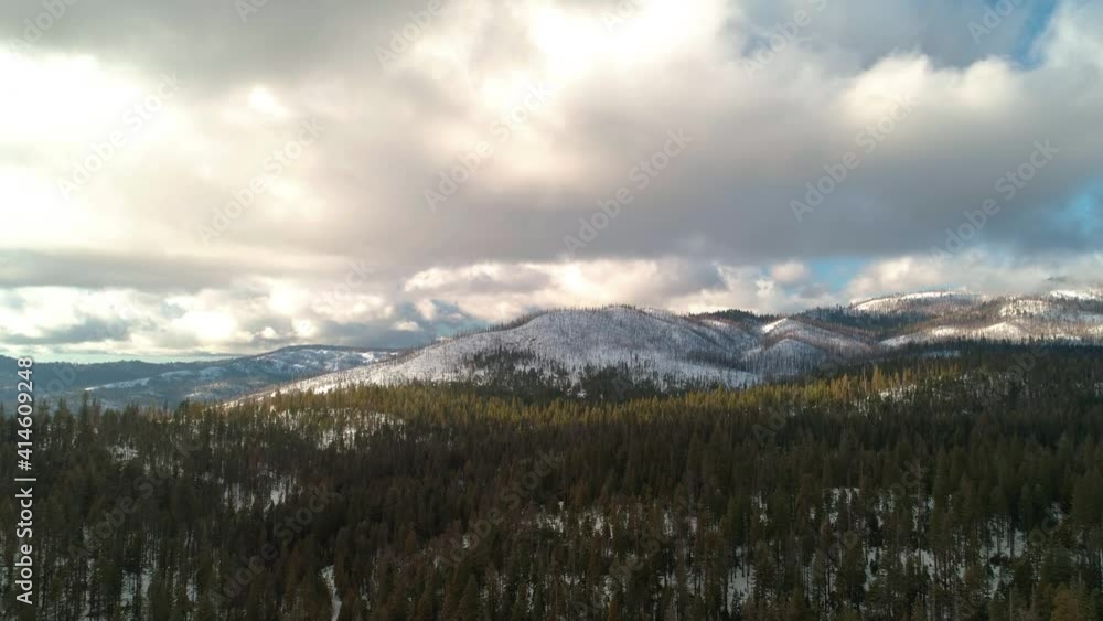 Epic aerial flying sideways over a forest with snow and mountains in the distance. Beautiful cloud formations, hilly terrain, lush green trees. Epic aerial flying sideways over a forest with snow and mountains in the distance. Beautiful cloud formations, hilly terrain, lush green trees.