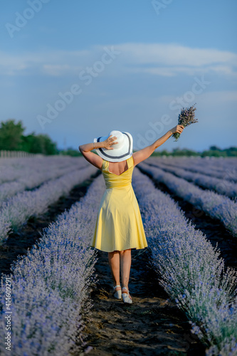Charming young woman in Blooming Lavender fields. Back view of lovely lady wearing waving yellow Dress, hat. Lavender essential oil with fresh lavender flowers, aromatherapy spa massage concept.