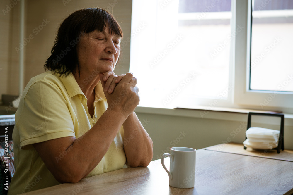 A mature woman sits at a table in the kitchen by the window with a pensive look A mature woman sits at a table in the kitchen by the window with a pensive look