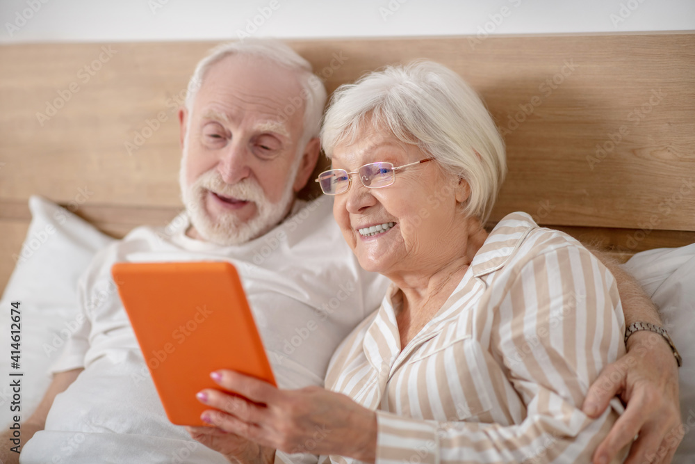 Aged married couple lying in bed and having good time while reading Aged married couple lying in bed and having good time while reading