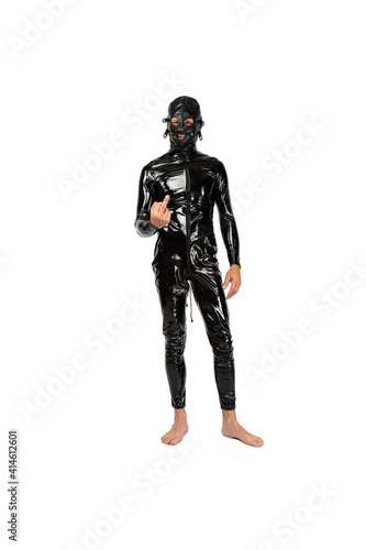 Man stands, dressed in latex fetish clothes and pvc mask showing middle finger, offensive gesture. bdsm costume for role-playing adult games. slave in the black catsuit looks directly into the camera