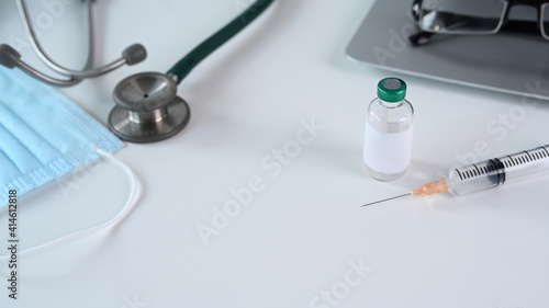 Doctor workplace with laptop, medical mask, stethoscope , vaccine bottle and syringes on white table.