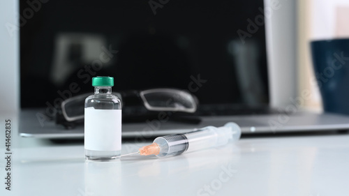 Close up view of vaccine bottle and syringes, laptop and glasses on doctor workplace.