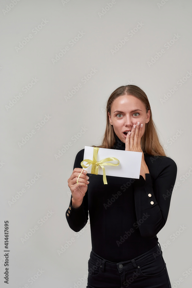 Young caucasian woman wearing black outfit looking shocked at camera, covering her mouth with hand, holding gift certificate for shopping while posing isolated over gray background Young caucasian woman wearing black outfit looking shocked at camera, covering her mouth with hand, holding gift certificate for shopping while posing isolated over gray background