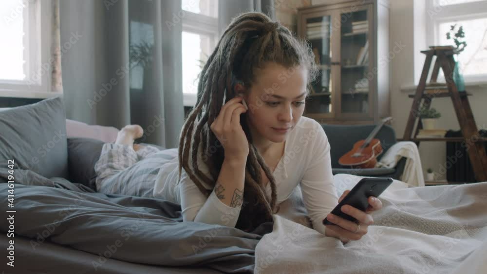 Slow-motion medium shot of smiling pretty woman with dreadlocks lying on bed in wireless headphones using smartphone in cozy relaxed atmosphere Slow-motion medium shot of smiling pretty woman with dreadlocks lying on bed in wireless headphones using smartphone in cozy relaxed atmosphere