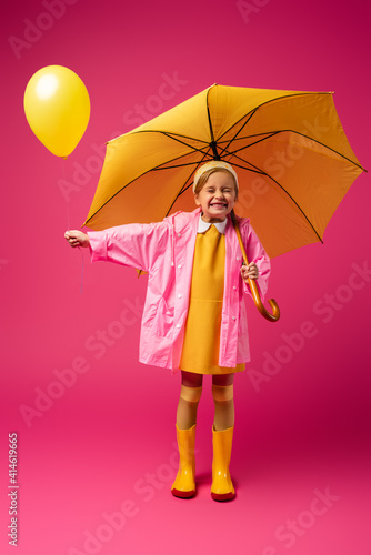 full length of excited girl in raincoat holding balloon and yellow umbrella on crimson © LIGHTFIELD STUDIOS