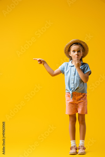 full length of shocked kid in straw hat covering mouth and pointing with finger on yellow