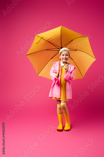 full length of happy girl in raincoat standing under yellow umbrella on crimson