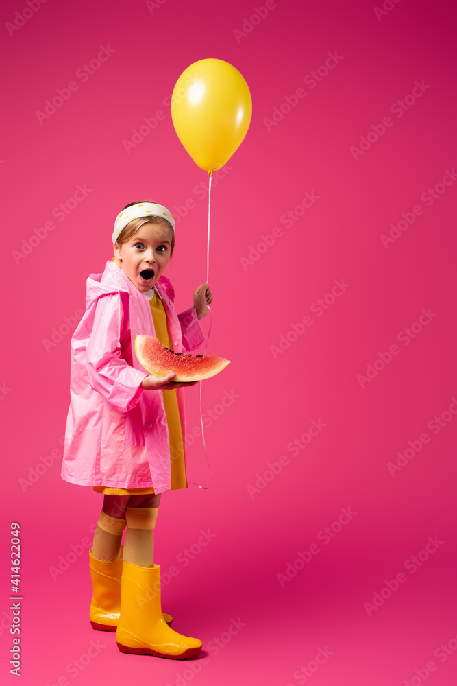 full length of amazed girl in raincoat holding balloon and sliced watermelon on crimson full length of amazed girl in raincoat holding balloon and sliced watermelon on crimson
