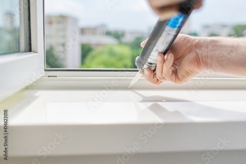 A worker with a construction syringe fills seam between sill and window with silicone sealant photo