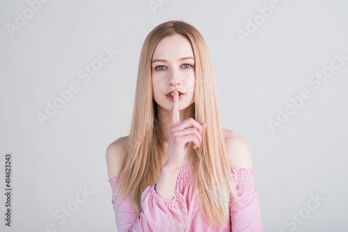 Portrait of a young blonde who holds her finger to her mouth in the studio on a white background. Secret!