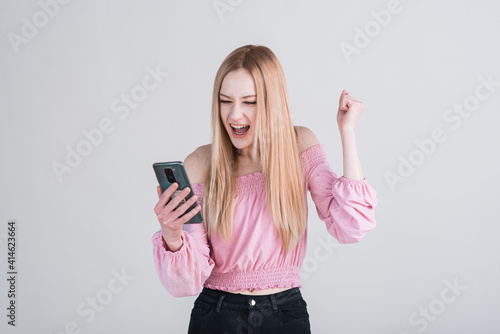 Portrait of a blonde girl who holds a smartphone and shows the gesture of the winner in the studio on a white background