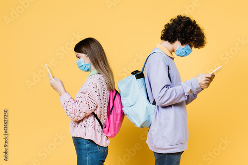 Side view of teen friends with backpacks and medical masks using digital tablets on yellow background