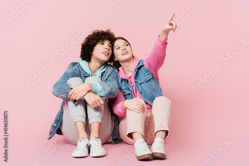 Smiling teen girl pointing with finger near boyfriend on pink background