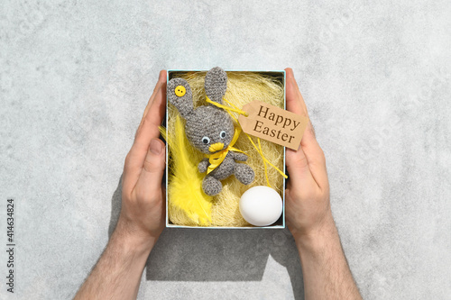 DIY Easter present making theme. Handmade knitted toy Easter rabbit in gift box, and male hands on grey background. Overhead view, flat lay