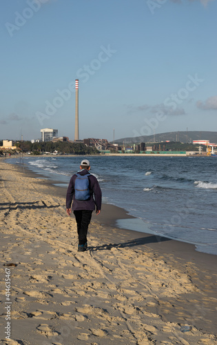 man walking alone on the beach with backpack and cap near an industry and fishing rods