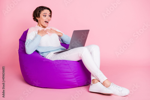 Full length photo portrait of amazed woman with laptop sitting in violet beanbag chair isolated on pastel pink colored background