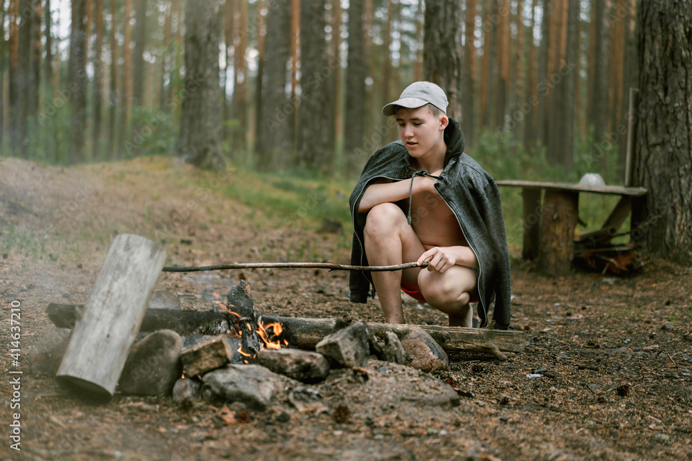 Caucasian teenage boy wearing a hoody jacket and cap, sitting by campfire in the forest holding long stick in hand. Image with selective focus Caucasian teenage boy wearing a hoody jacket and cap, sitting by campfire in the forest holding long stick in hand. Image with selective focus